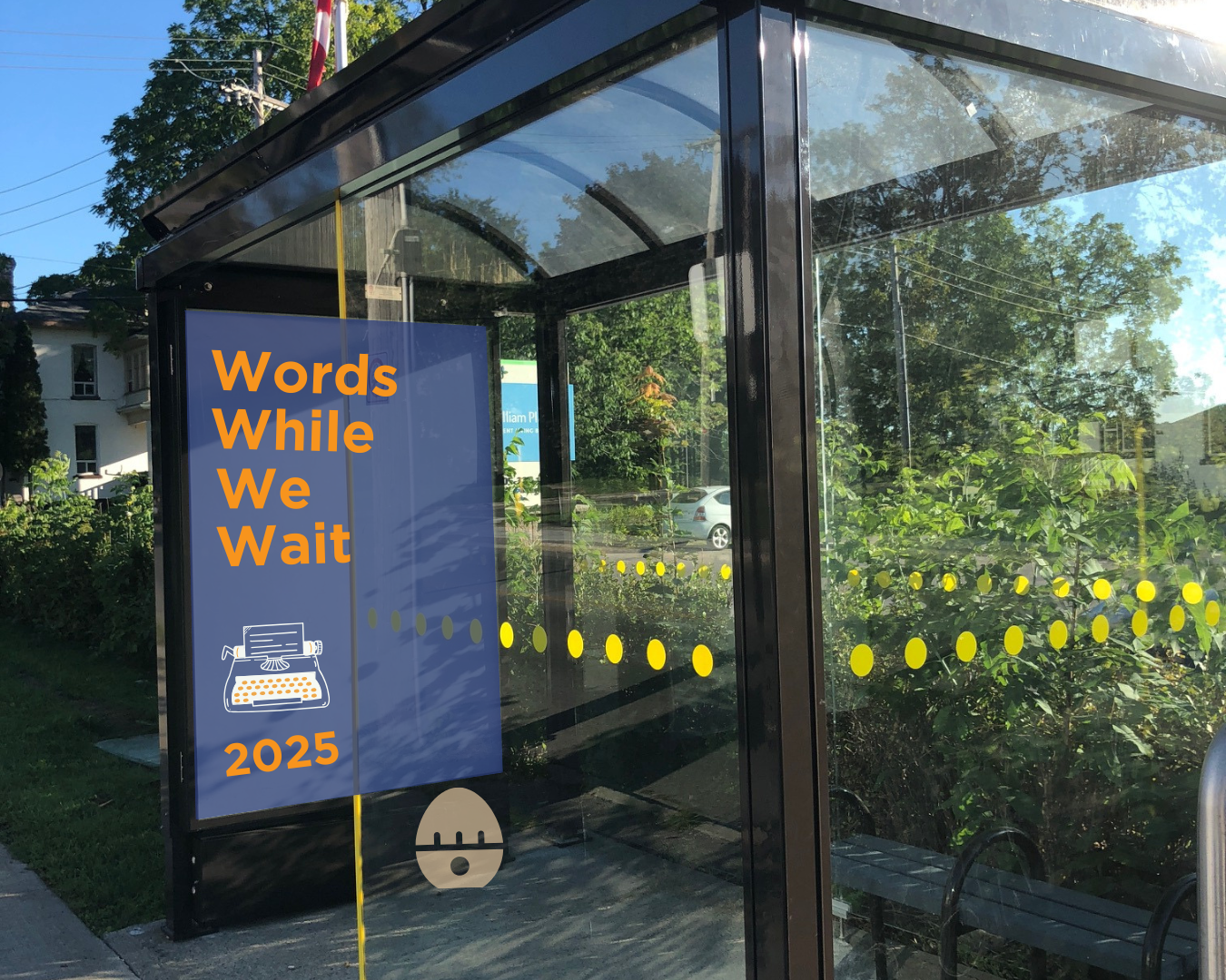Bus shelter with a blue poster displaying the words &quot;Words While We Wait 2025.&quot; Also on the poster is a typewriter graphic.
