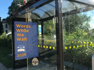 Bus shelter with a blue poster displaying the words &quot;Words While We Wait 2025.&quot; Also on the poster is a typewriter graphic.