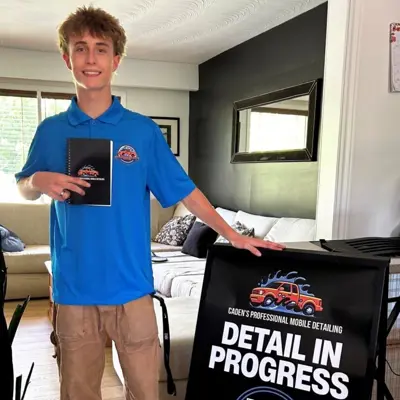 A young man is standing and smiling while touching a sign that says 'Detail in Progress'.