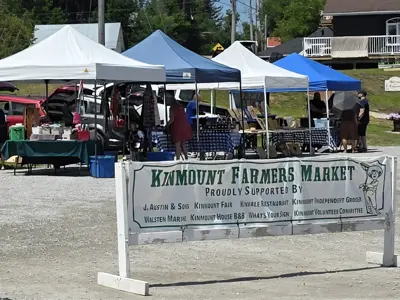 Farmers' market tents with products and customers in the background and a Kinmount Farmers' Market banner in the foreground.