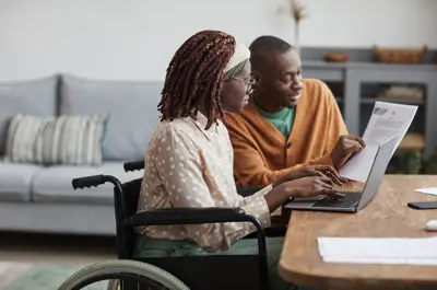 Two people sitting at table looking at laptop and piece of paper.
