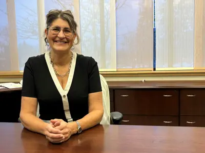 Woman wearing glasses sits at a table. She is smiling, and trees are visible through the windows in the background.