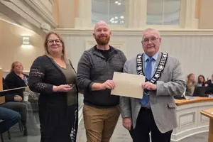 Photo of three people standing and facing the camera. The centre person is holding an envelope.