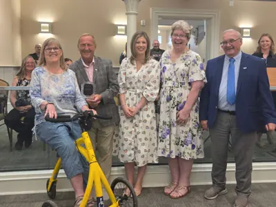 Photo of a group of people posed as one holds an award. One woman is seated on a yellow mobility trike.