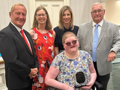 A young woman using a wheelchair sits holding an award. Two men and two women stand behind her.