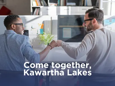 Photo of two male colleagues wearing glasses, one black and one white, giving a fistbump. They are seated at a desk and there is a text overlay reading 'Come together, Kawarth Lakes.'