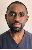 Portrait photo of a man with short hair and dark skin tone looking directly at the camera. He is wearing a dark blue scrub shirt.
