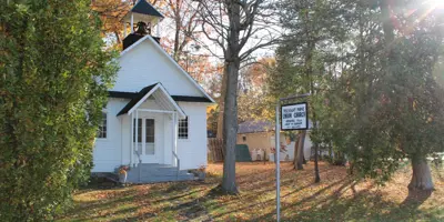 Photo of a white clapboard church with a steel bell in the small steeple. A sign in front reads 'Pleasant Point Union Church.' The ground around the church is grassy and dotted with orange and brown leaves that have fallen.