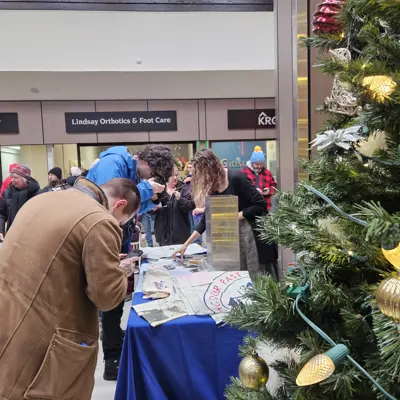 Photo of people looking at items displayed on a table. A decorated Christmas tree is in the foreground.