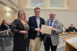 Photo of three people standing and facing the camera. The centre person is holding an envelope.