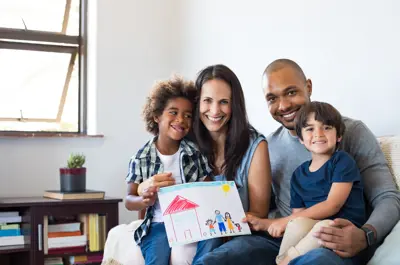 Family with two children, a man and a woman smiling and sitting on a coach holding up a child's drawing
