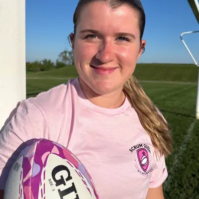 A young woman in a pink shirt is smiling while facing the camera and holding a rugby ball.