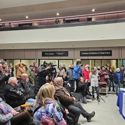 Photo of people sitting and standing in an audience, facing a table.