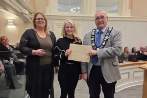 Photo of three people standing and facing the camera. The centre person is holding an envelope.