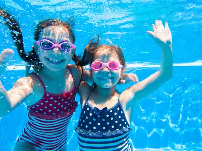 two campers smiling underwater and waving at the camera during their aqua fun camp