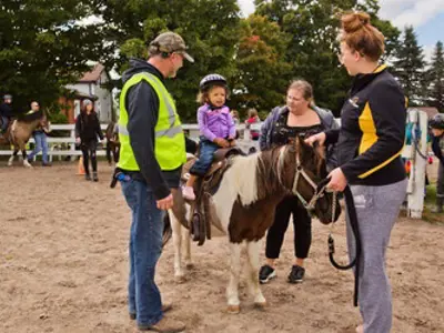 Child wearing a helmet riding a pony with a leader, and safety person assisting, and parent walking beside.