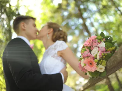 A bride and groom kiss in the background, with focus on the bride's floral bouquet