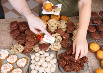 A variety of baked goods on a table. Two hands are reaching for goodies, and a tag reads 'Bake Sale'