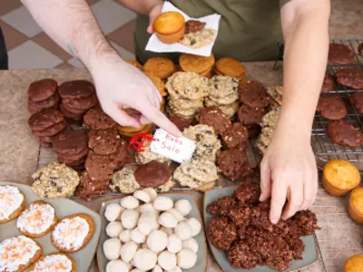 A variety of baked goods on a table. Two hands are reaching for goodies, and a tag reads 'Bake Sale'