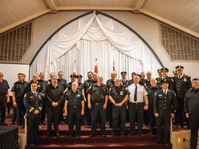 A large group of uniformed firefighters stand on an indoor stage in front of a curtained backdrop