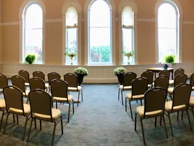 Room with rows of chairs with an aisle facing tall windows and flowers