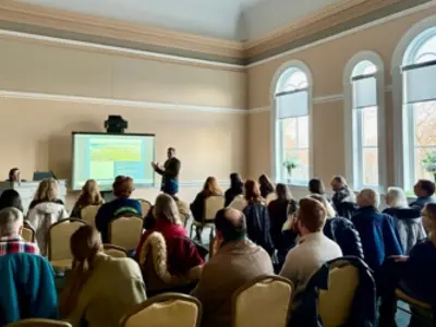 A group of adults sit on chairs and watch a speaker making a presentation in front of a screen.