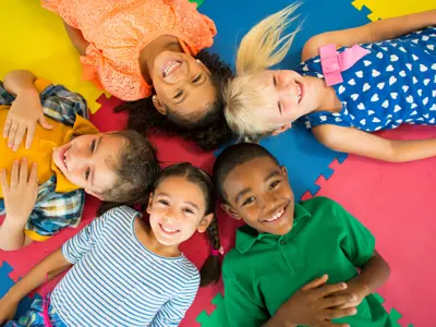 Photo of five children of varied skin and hair colours lying on the floor with their heads together. They are all smiling.