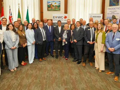 A large group of men and women in businesswear pose for a photo in front of flags and signs.