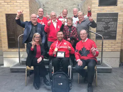 Members of Council pose with a man wearing a gold medal. He sits in a wheelchair, and the councillors surround him and hold up one finger to represent 'number one'. Many are wearing red in celebration of a Canadian gold medal.