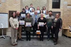 A group of six people are smiling and standing indoors on a staircase. The background is yellow brick.
