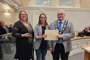 Photo of three people standing and facing the camera. The centre person is holding an envelope.