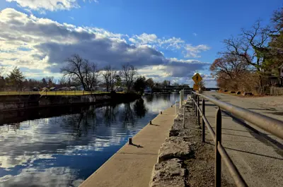 A canal beside a paved path with a railing, leafless trees, and a pedestrian sign under a partly cloudy sky.