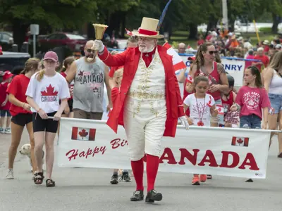 Man in Canada attire walking in front of banner that reads Happy Birthday Canada with many adults and children walking behind him