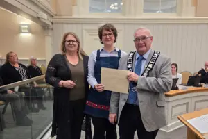 Photo of three people standing and facing the camera. The centre person is holding an envelope.