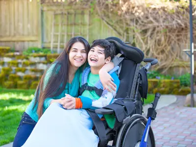 A woman embraces a young boy sitting in a wheelchair; both smile at the camera