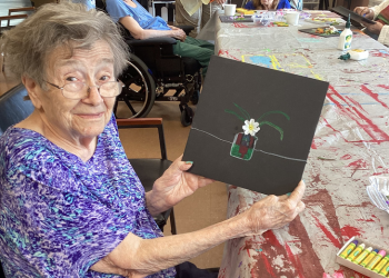 An older woman wearing glasses is smiling and holding her mixed-media artwork.