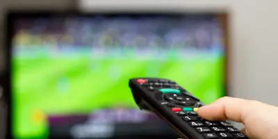 Hand holding remote control with TV on in the background.