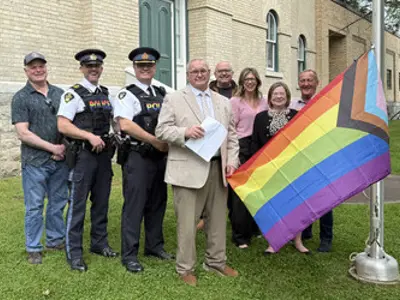 Officials and uniformed police officers standing near a flag pole. One man holds the rainbow Pride flag that is attached to the pole and about to be raised.
