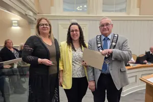 Photo of three people standing and facing the camera. The centre person is holding an envelope.