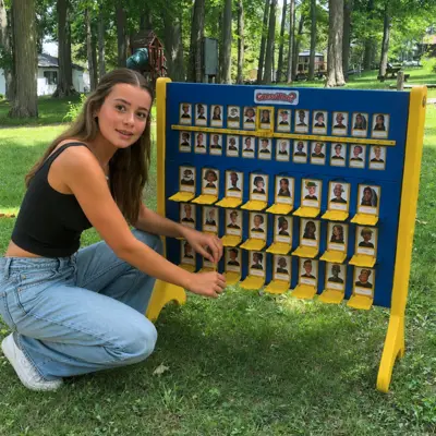 A young woman is crouched down beside a lawn game on the grass, with trees in the background.