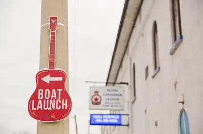 Pole with a red guitar cut out that reads Boat Launch with an arrow pointing left. White building with business sign is in the background.