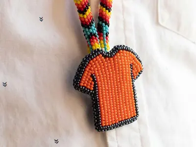 Photo of an Indigenous beaded necklace, with a beaded orange shirt pendant. A white button-up shirt is the background.
