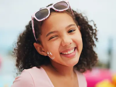 camper smiling and winkig at the camera with sunglases on their head and smiley face on their cheek made with sunscreen