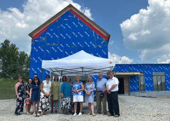 A group of officials stand in front of a building under construction. The building is wrapped in a blue construction material.