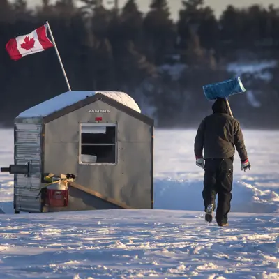 A person with a shovel walks toward an ice fishing hut on a frozen lake