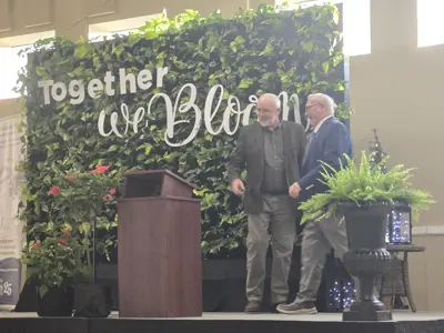 Two men stand on a stage with a podium nearby and in front of a living wall that reads "Together We Bloom"