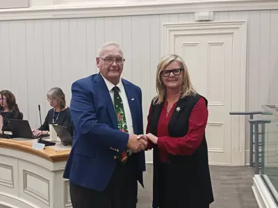 A man wearing a blue blazer shakes hands with a woman wearing a red sweater and black vest. They are smiling for the camera.