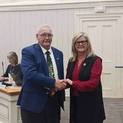 A man wearing a blue blazer shakes hands with a woman wearing a red sweater and black vest. They are smiling for the camera.