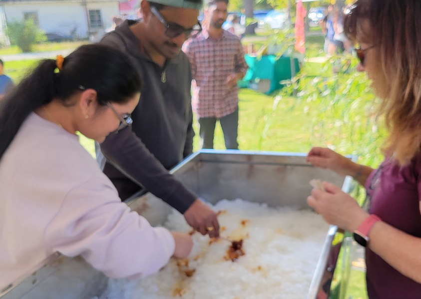 Woman rolling maple taffy onto a popsicle stick in a tub of crushed ice. A man is doing the same while another woman guides them.