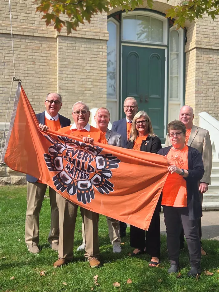 Five men and two women stand behind an orange flag they are holding. The flag has text reading 'Every Child Matters.'